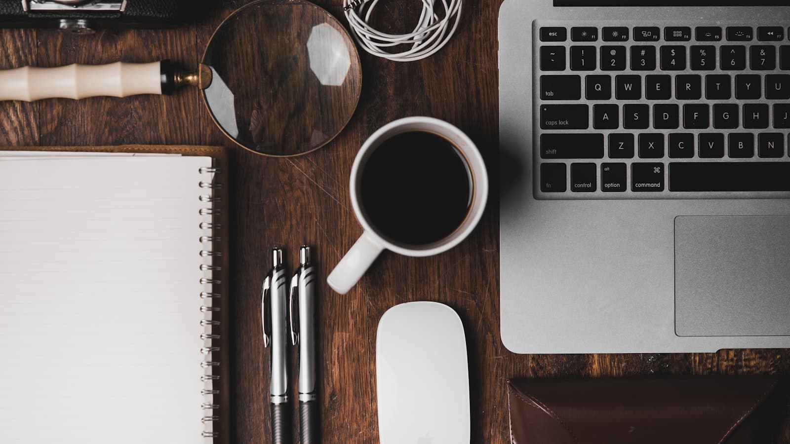 Flat lay of a notebook, pens, a magnifying glass and a coffee cup beside a laptop keyboard