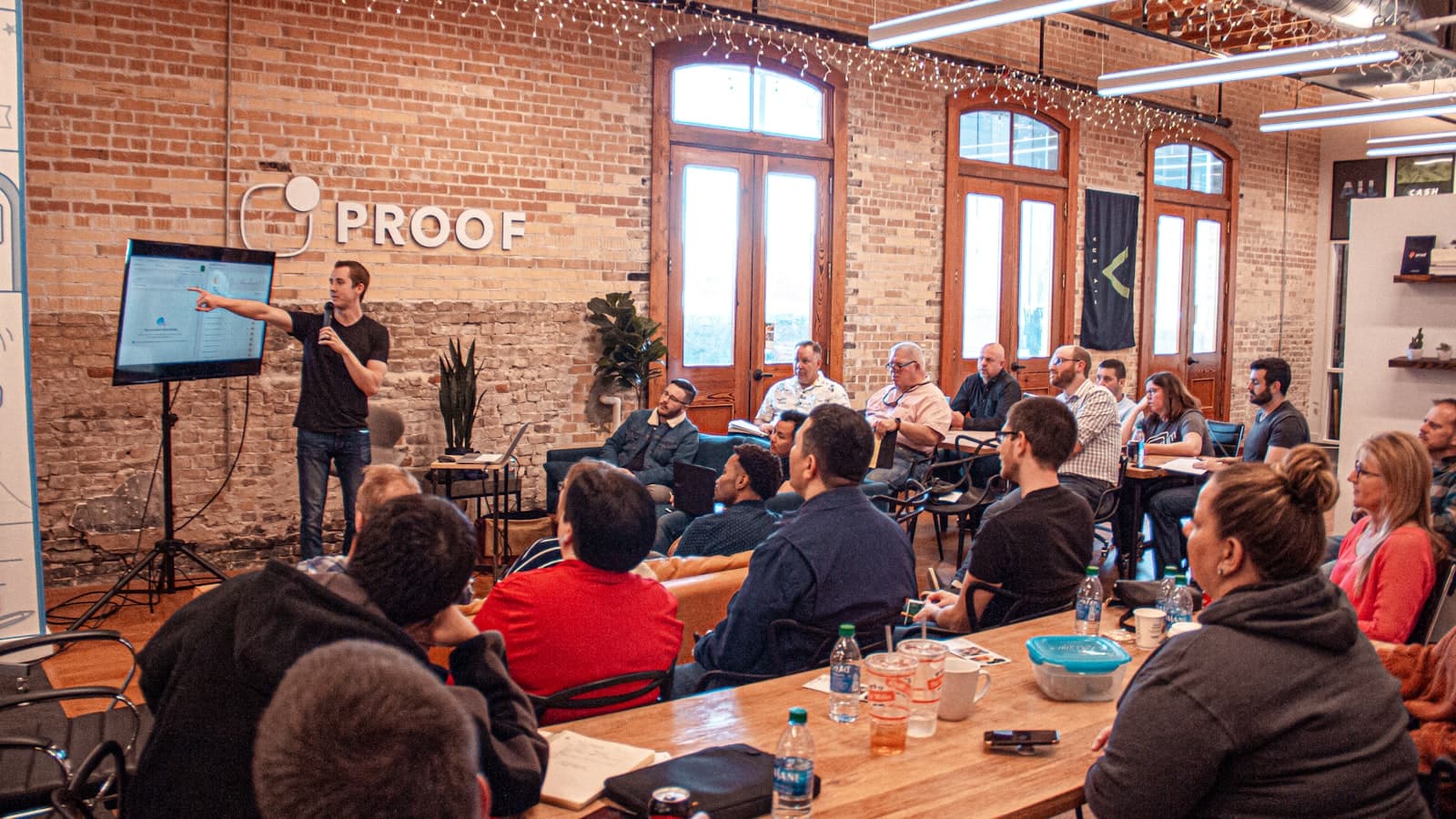 Speaker presenting to a seated audience in a brick-walled room, with people listening attentively